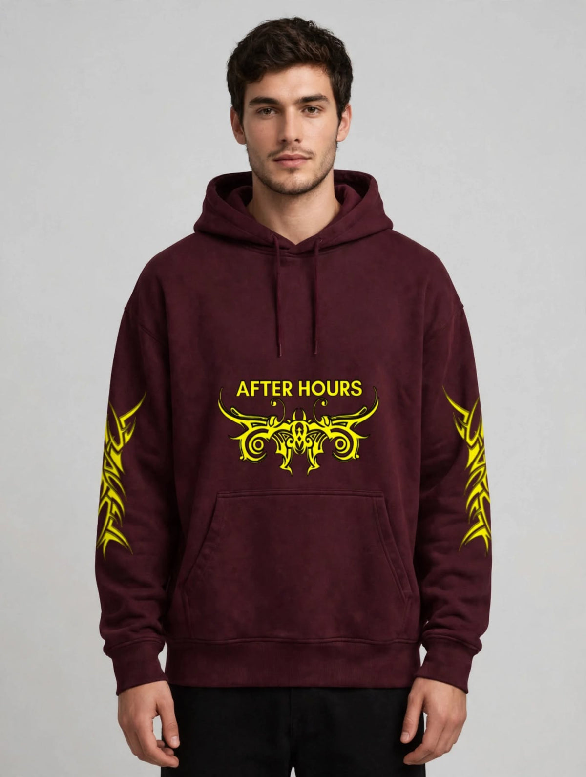 A man wearing a maroon pullover hoodie with yellow graphic prints on the chest and sleeves, standing against a light grey studio background.
