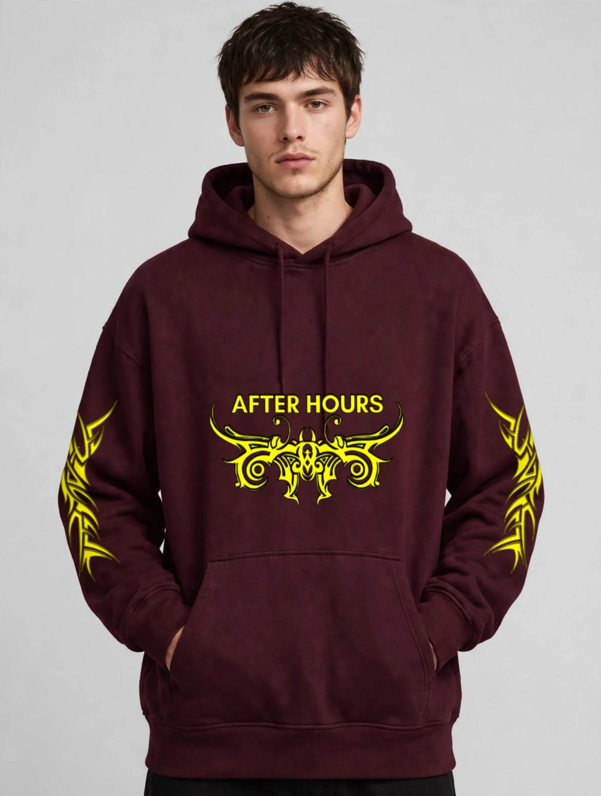 A man wearing a maroon pullover hoodie with a yellow graphic print on the chest reading 'AFTER HOURS' and matching yellow tribal-style sleeve prints, photographed against a light grey studio background.