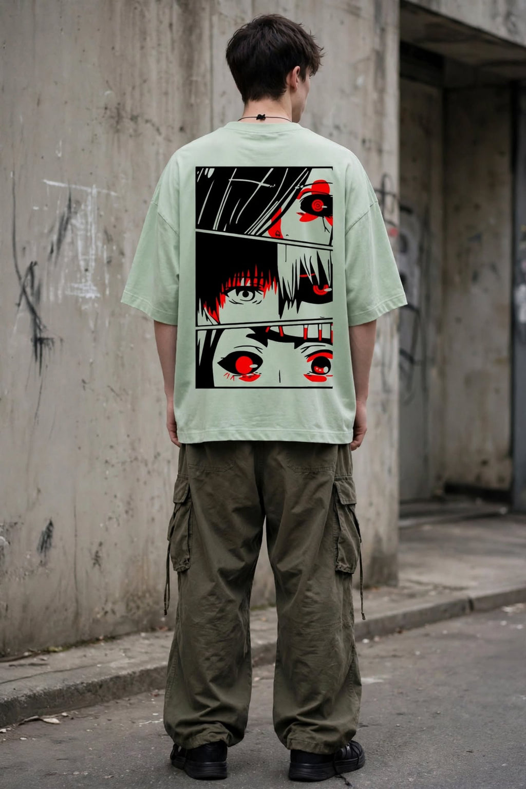 A person standing outdoors against a concrete wall, viewed from behind, wearing an oversized light sage green T-shirt with a large black, white, and red anime-style graphic print on the back, paired with olive cargo pants and black shoes.