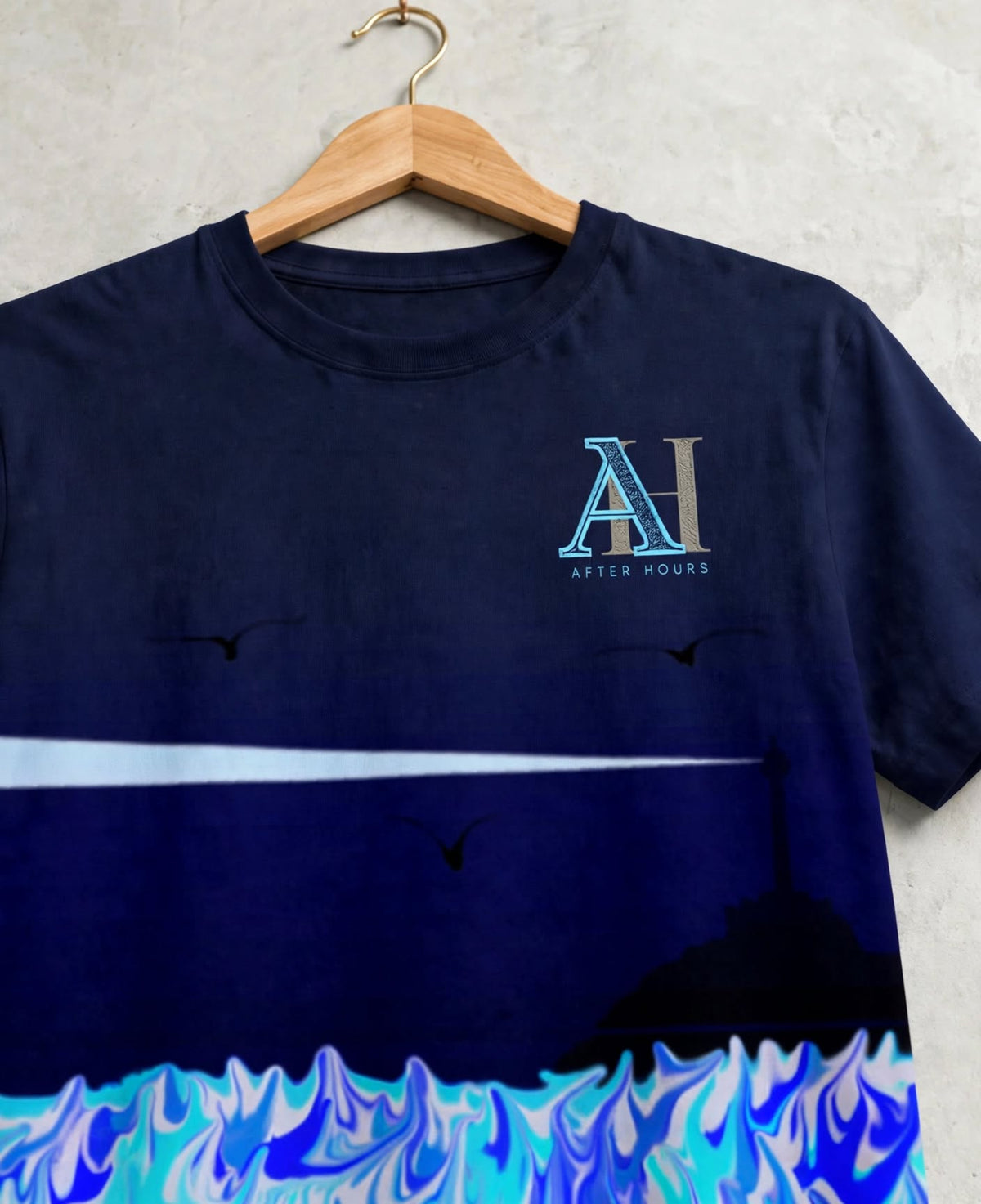 A navy blue graphic t-shirt hanging on a wooden hanger against a textured light gray wall. The shirt features a stylized 'AH' logo with 'AFTER HOURS' text on the left chest, a white horizontal light streak from a lighthouse across the middle, and a blue and white marbled wave design along the bottom hem.