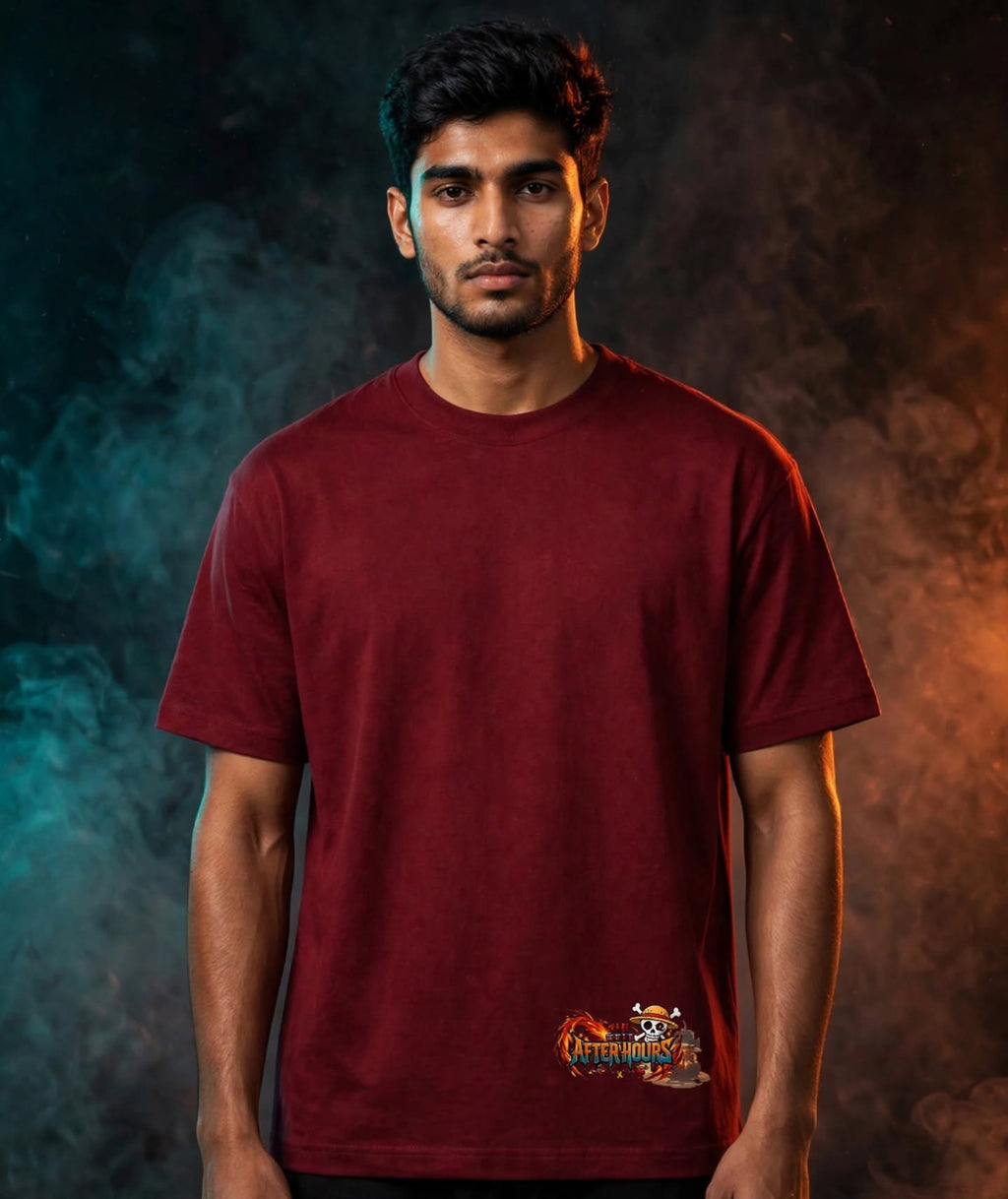 A man wearing a maroon short-sleeve t-shirt with a small cartoon graphic on the bottom left, set against a dark smoky background with blue and orange lighting.