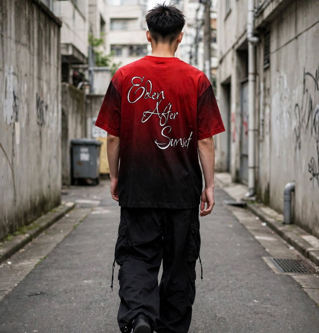 A man viewed from behind walking down a narrow urban alley wearing a red-to-black gradient t-shirt with 'Eden After Sunset' written in white script across the back, paired with black cargo pants.