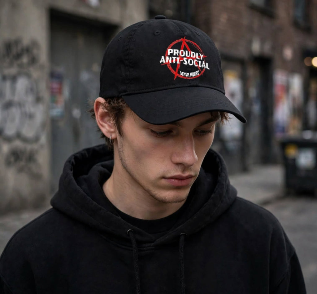 A man wearing a black baseball cap with a red and white 'Proudly Anti Social' graphic, paired with a black hoodie, standing in an urban alleyway.