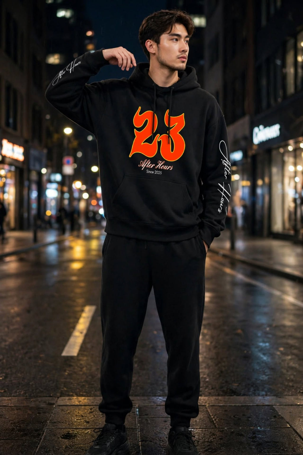 A man stands on a wet city street at night wearing a black hoodie with a large orange '23' graphic and white script text on the front, paired with black sweatpants and black shoes.