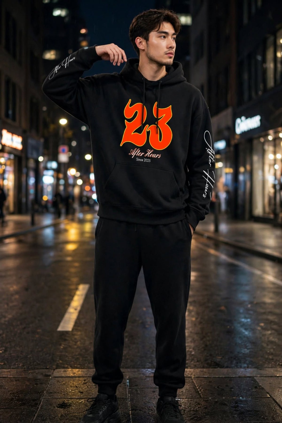 A man stands on a wet city street at night wearing a black hoodie with a large orange '23' graphic and white script text on the front, paired with black sweatpants and black shoes.