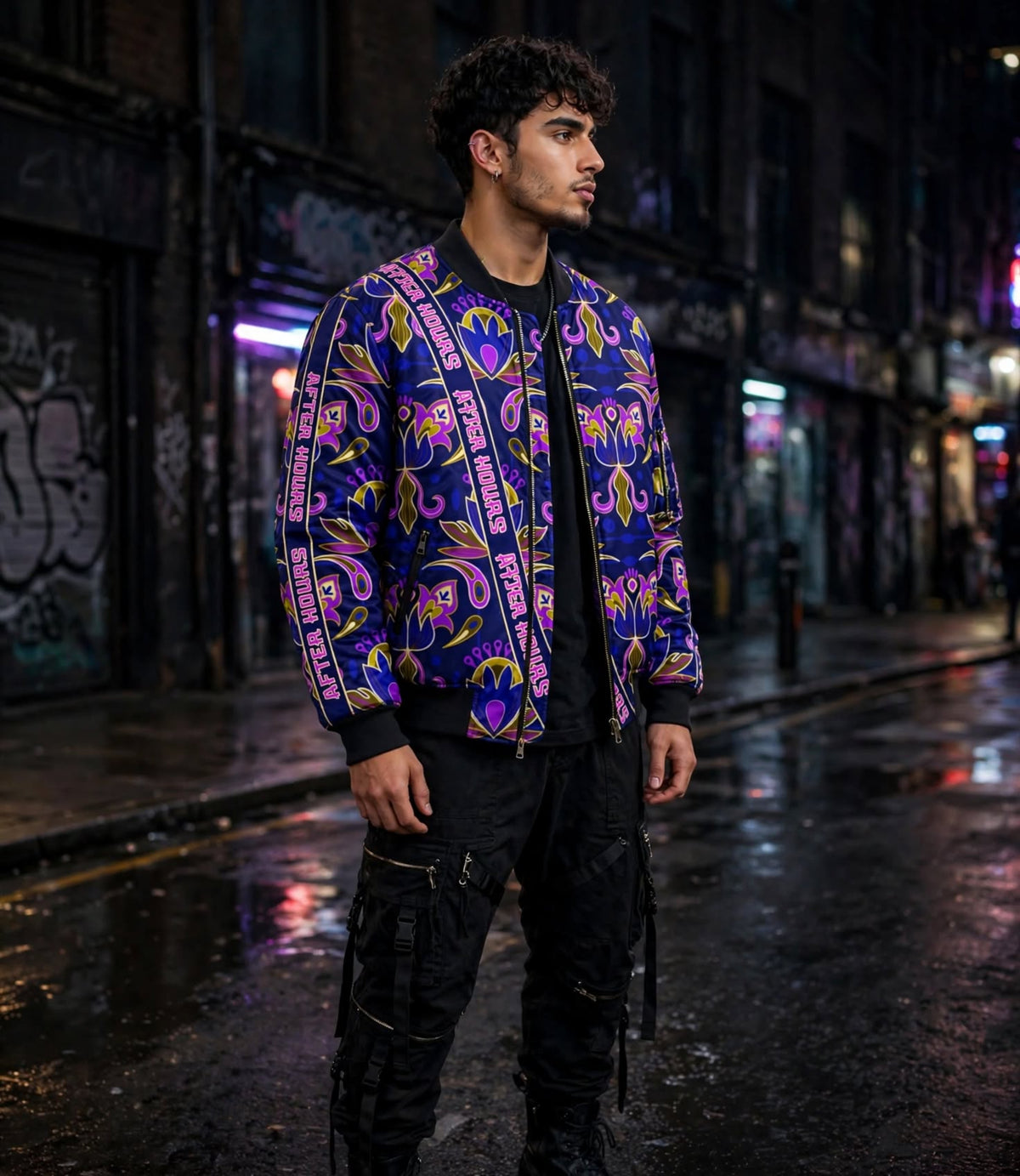 A man wearing a vibrant purple and pink paisley bomber jacket with 'AFTER HOURS' printed down the sleeves, standing on a wet city street at night.