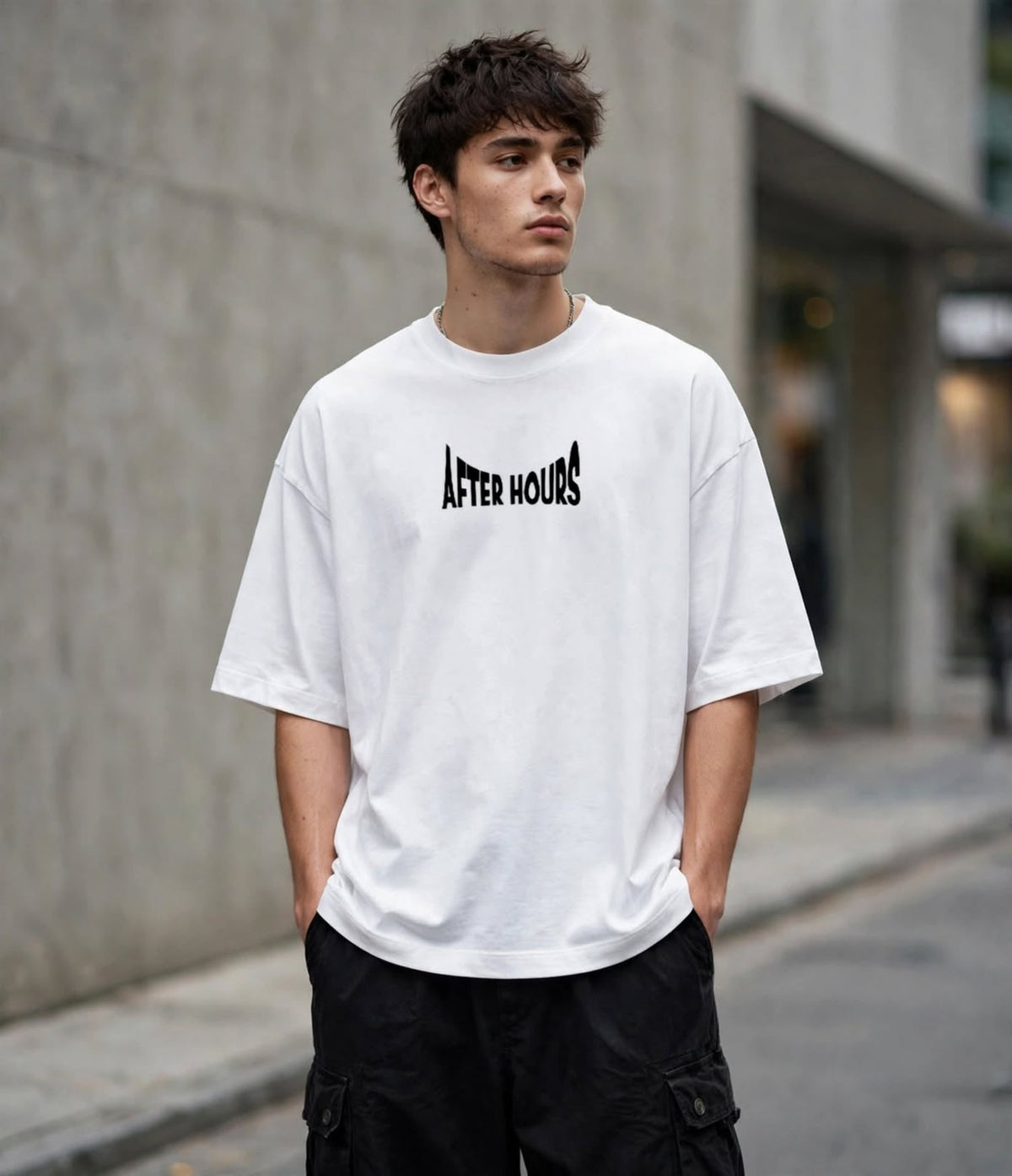 A man wearing a white oversized t-shirt with a black 'AFTER HOURS' graphic on the chest, paired with black cargo pants, standing outdoors against a blurred concrete wall.