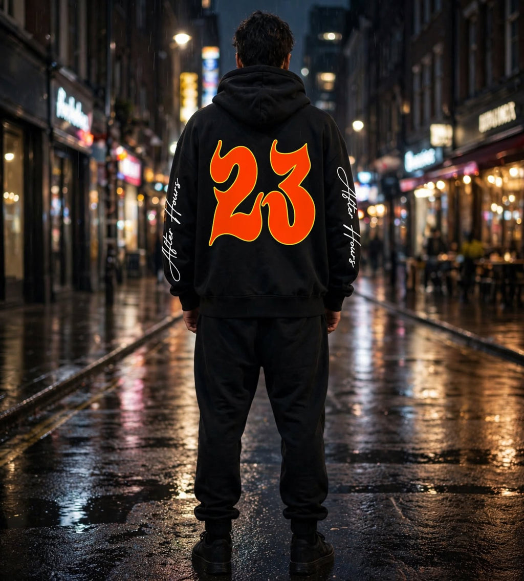 A person standing on a wet city street at night, viewed from behind, wearing a black hoodie with a large orange '23' graphic on the back and white script text down both sleeves.