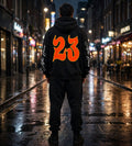A person standing on a wet city street at night, viewed from behind, wearing a black hoodie with a large orange '23' graphic on the back and white script text down both sleeves.