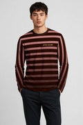 A man wearing a long-sleeve striped knit top with dark brown and dusty rose horizontal stripes, featuring the 'AFTER HOURS' logo on the chest, paired with dark trousers, against a light grey studio background.