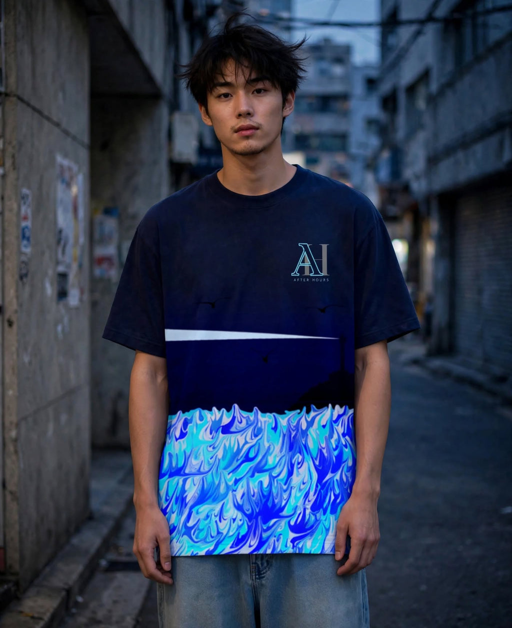 A young man stands in a narrow urban alley wearing a short-sleeve graphic T-shirt. The shirt is dark navy at the top with a white horizontal stripe across the chest and a vivid blue, cyan, and light blue wave-like pattern across the lower half.