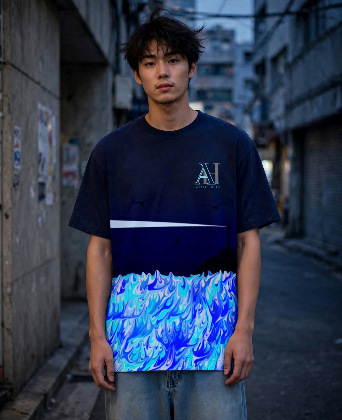 A young man stands in a narrow urban alley wearing a short-sleeve graphic T-shirt. The shirt is dark navy at the top with a white horizontal stripe across the chest and a vivid blue, cyan, and light blue wave-like pattern across the lower half.