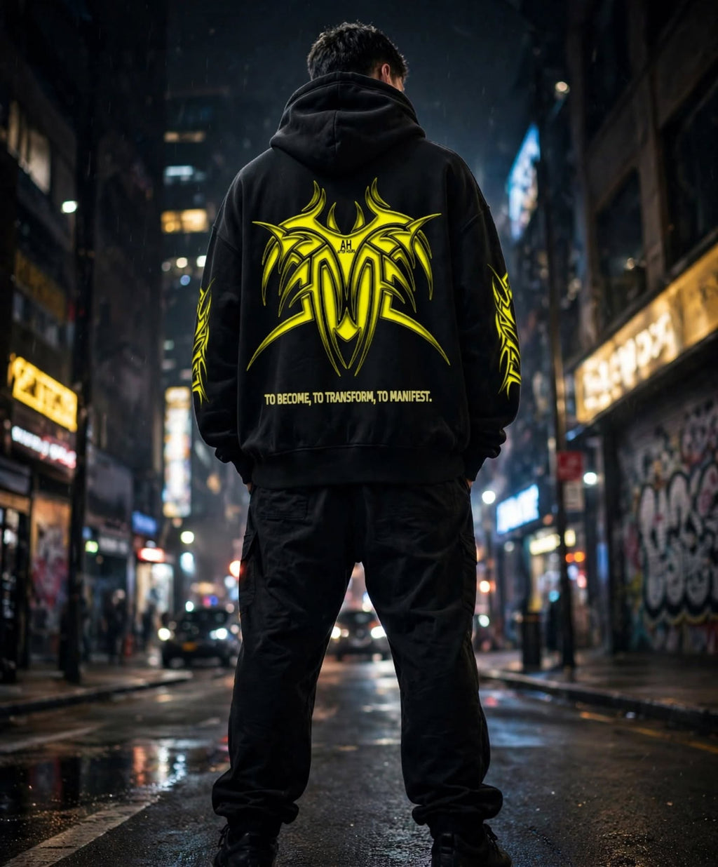 A man stands on a wet city street at night, viewed from behind, wearing a black hoodie with a large bright yellow graphic and text on the back, paired with black pants.