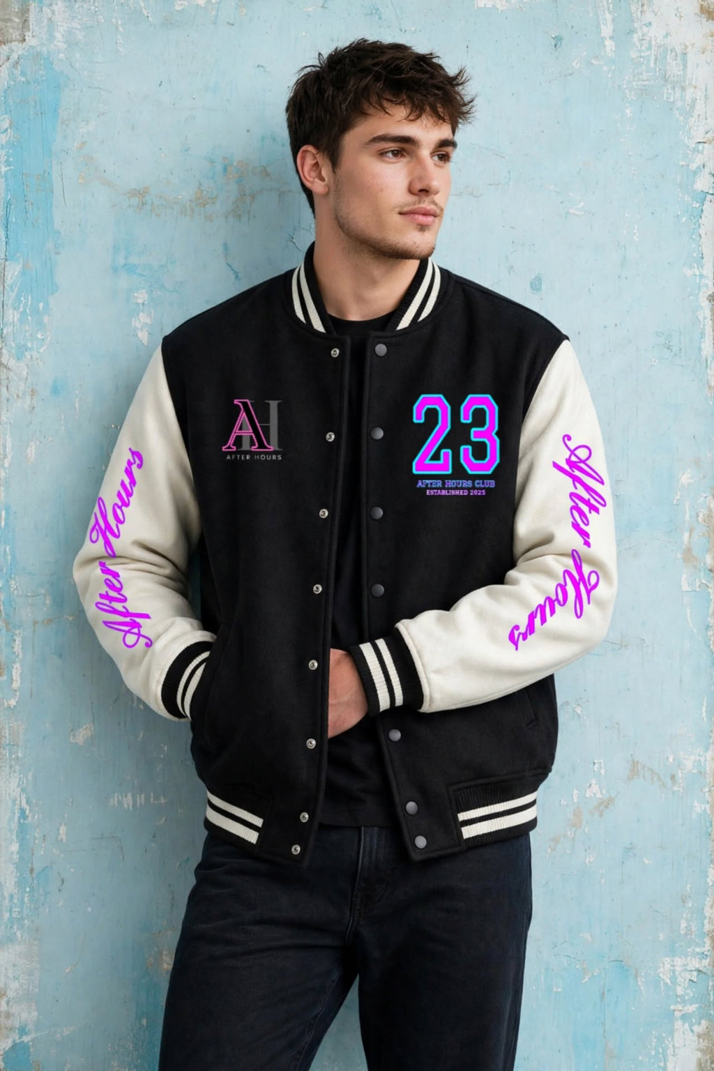 A man wearing a black and cream varsity jacket with pink and blue lettering, posing against a textured light blue wall.